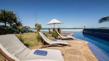Zona de piscina con sillones reclinables y una sombrilla, con vista al océano.