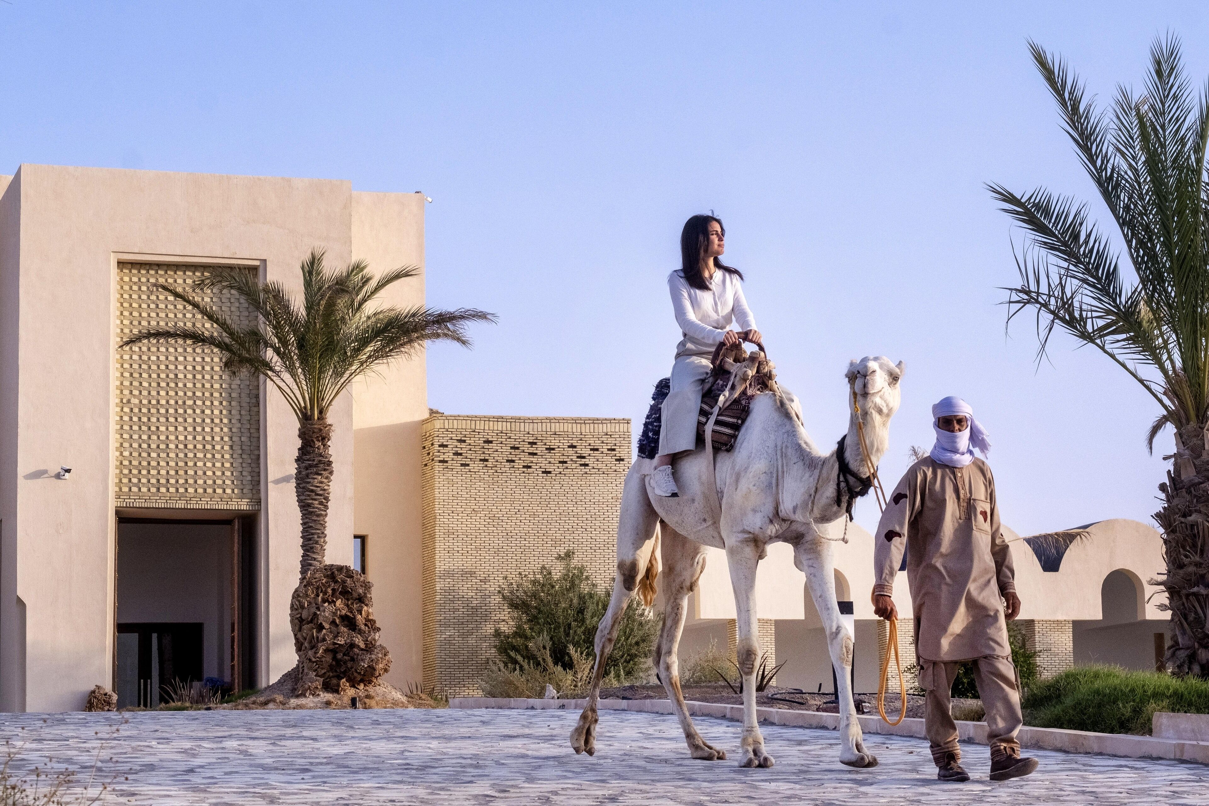 A person riding a camel, a man walking beside them, and a building with a patterned facade.