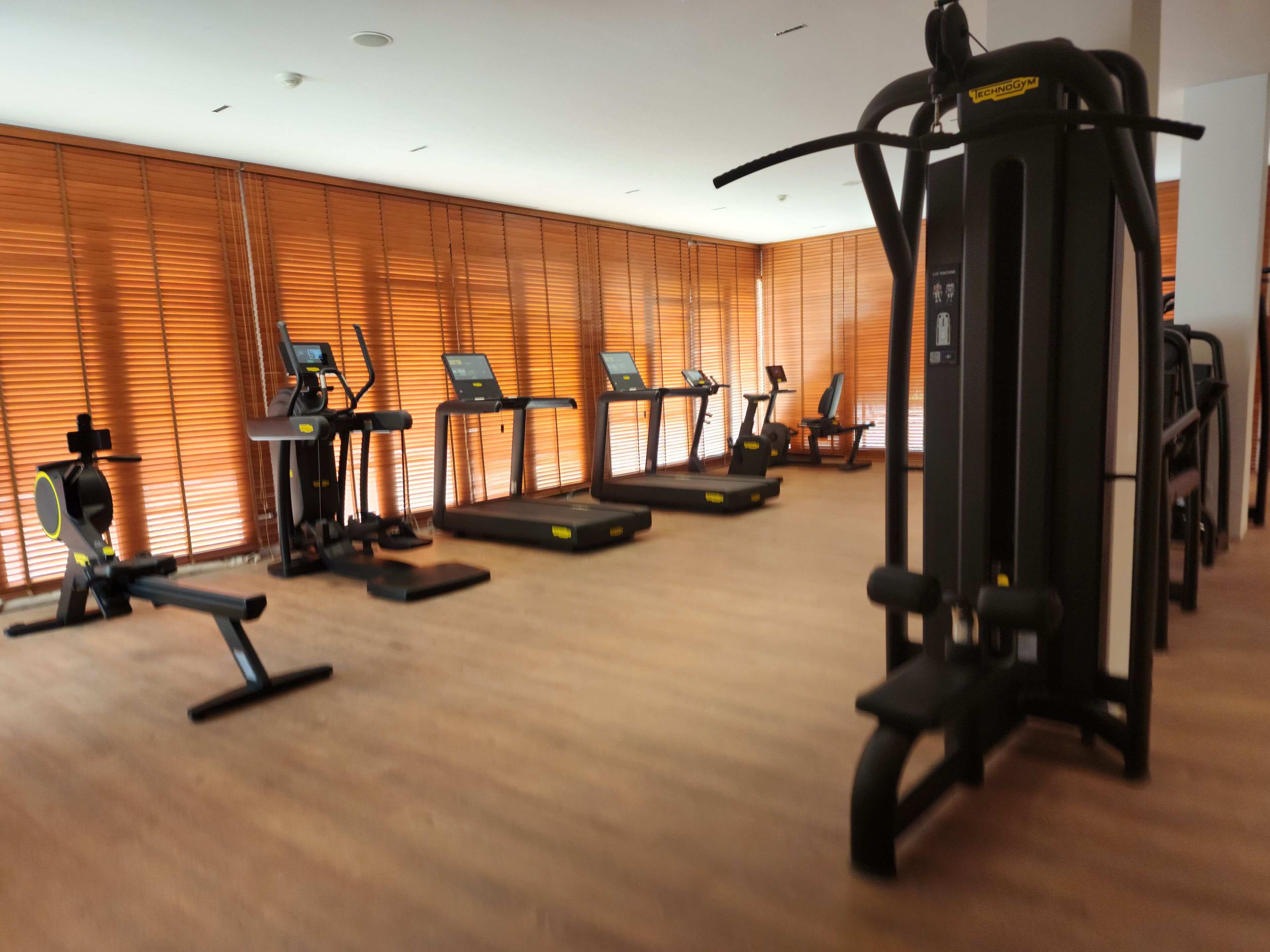 Une salle de sport équipée de divers appareils d’exercice, dotée de planchers en bois et de grandes fenêtres avec stores.