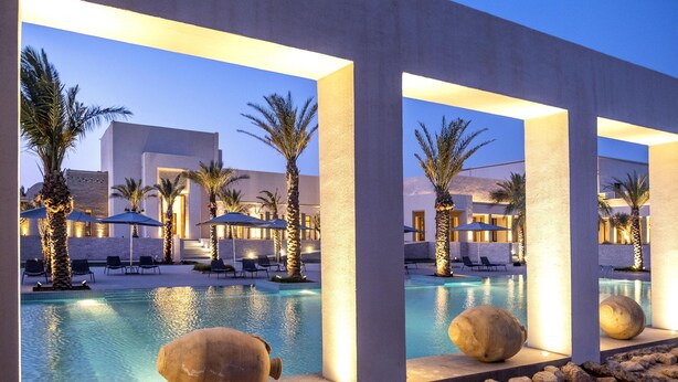 Uma área de piscina moderna, com espreguiçadeiras e palmeiras, cercada por edifícios com iluminação acolhedora.