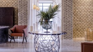 Um lobby com parede de tijolos, uma mesa moderna com base geométrica, uma poltrona de couro marrom e um vaso de plantas.