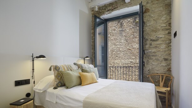 A bedroom with a bed, bedside lamps, a wooden nightstand, a chair, and a stone wall. - Canvas Apartments & Lofts Girona, Situados en el río Onyar (Girona)
