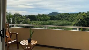 A balcony with a wooden chair and a small table, overlooking a lush green landscape with trees and hills. - Condo Marimba, near Sámara and Carrillo beach (Sámara)