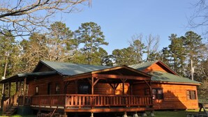 A wooden cabin with a porch and a green roof, surrounded by trees. - COZY CABIN/GREAT RATES!! Acreage to walk. Pond views. MAX 8 guests, No Pets (Broken Bow)