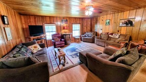 A cozy log cabin interior with a wooden ceiling, a television, leather armchairs, and a patterned rug. - COZY CABIN/GREAT RATES!! Acreage to walk. Pond views. MAX 8 guests, No Pets (Broken Bow)