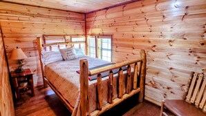 A wooden cabin bedroom with a bed, a chair, a lamp, and a window. - COZY CABIN/GREAT RATES!! Acreage to walk. Pond views. MAX 8 guests, No Pets (Broken Bow)