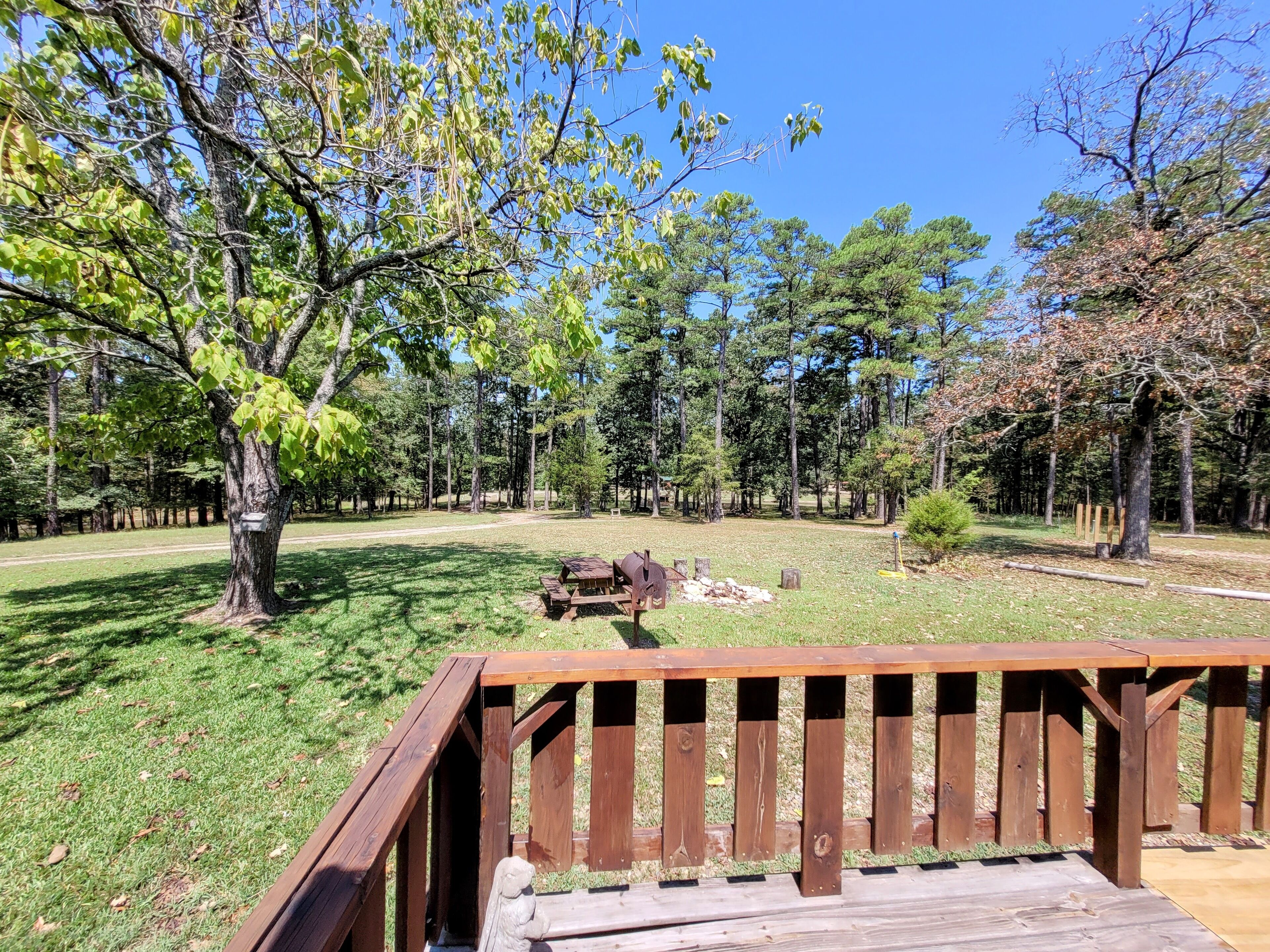 Une terrasse en bois avec une table de pique-nique et un banc, entourée d’une zone herbeuse bordée d’arbres.