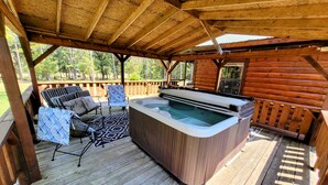 A wooden deck with a hot tub, outdoor furniture, and a view of the surrounding forest. - COZY CABIN/GREAT RATES!! Acreage to walk. Pond views. MAX 8 guests, No Pets (Broken Bow)