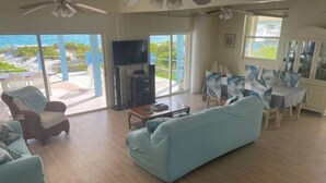 En rummelig stue med en stor lyseblå sofa, et træbord og et fjernsyn. Der er et spisebord med stole, og en balkon med udsigt til havet.