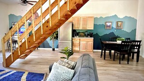 Un salon avec un canapé gris, un escalier en bois, une cuisine avec des meubles en bois et un coin repas avec des chaises.
