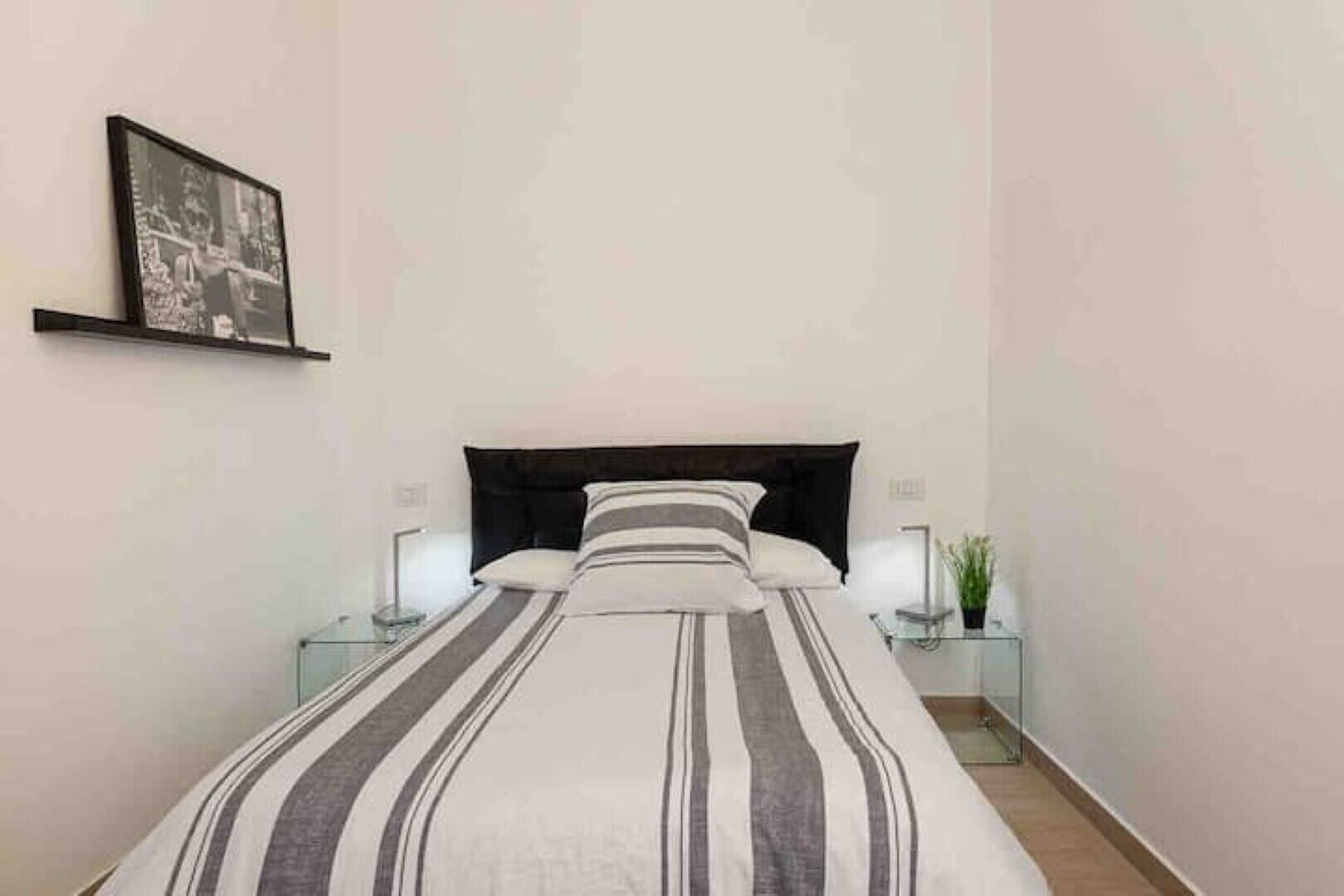 Un dormitorio ordenado con cama, dos mesitas de noche, una lámpara de noche, un cuadro en la pared y una planta en maceta.