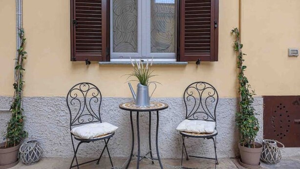 Zwei Metallstühle mit Kissen, ein kleiner runder Tisch mit einem Blumentopf und ein Fenster mit braunen Rollläden.