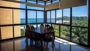 Un comedor con un ventanal grande con vistas a una playa y vegetación exuberante.