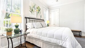 A bedroom with a bed, bedside tables, a window with blinds, and artwork on the wall. - Classic Coastal Cottage ~ Steps to the Beach! 4-Seater Golf cart for rent! (Bay St. Louis)