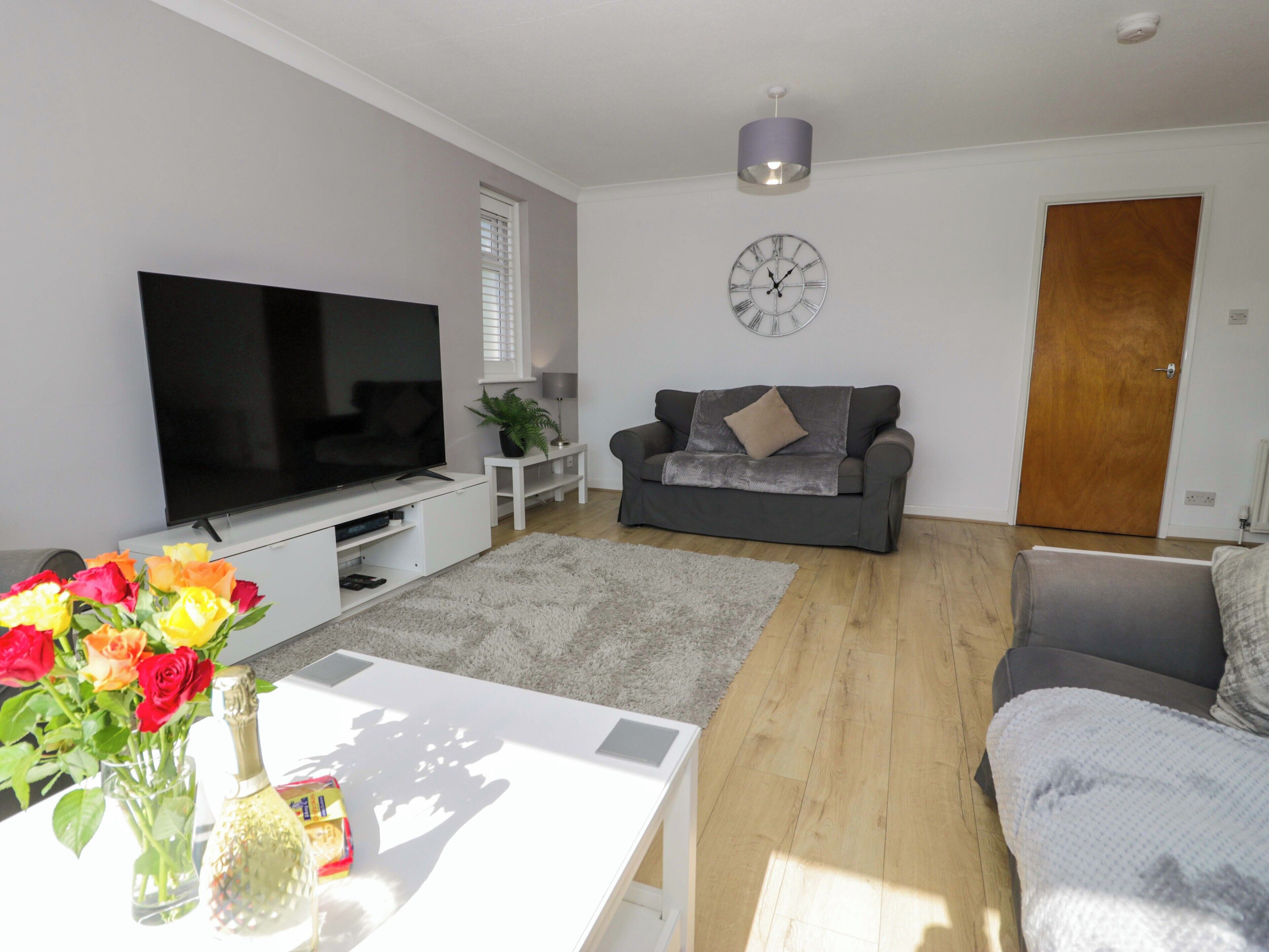 A living room with a flat-screen TV, a grey sofa, a white coffee table, and a wooden floor.