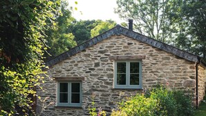 Una casa de piedra con dos ventanas y techo inclinado, rodeada de abundante vegetación y plantas con flores.