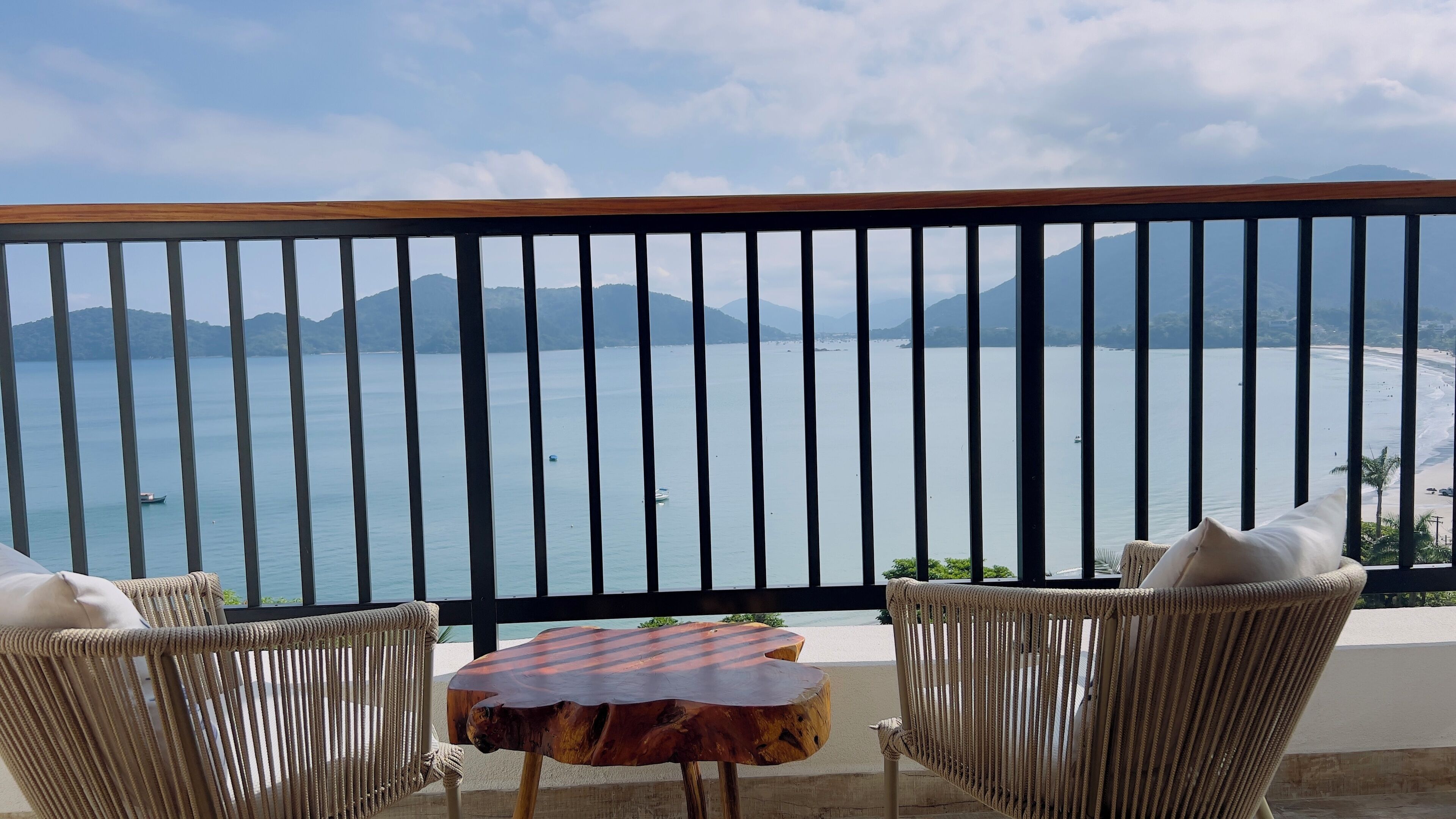 A balcony with wicker chairs and a small table, overlooking a calm sea with boats and distant mountains.