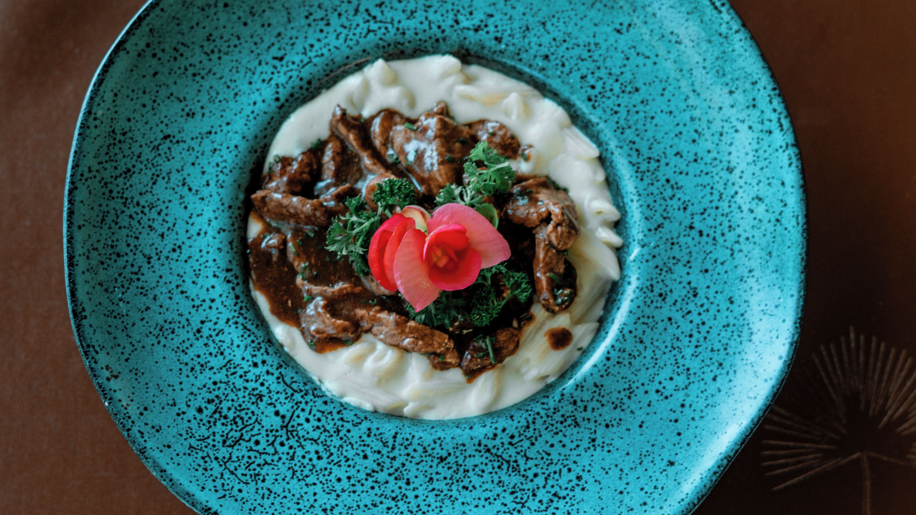 Un plato con guiso de carne y salsa, adornado con una flor, sobre un plato azul moteado.