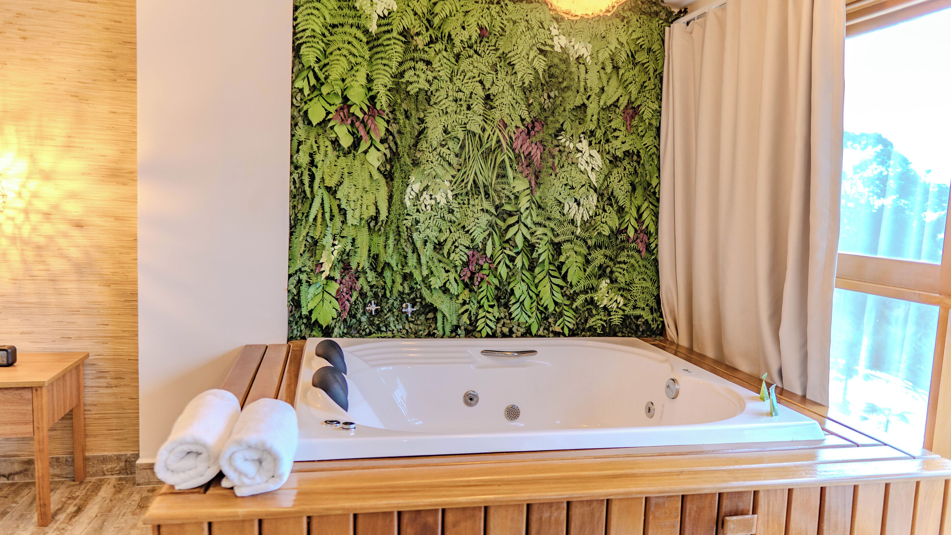 Una habitación con una bañera grande, suelo de madera y una pared decorada con follaje verde.