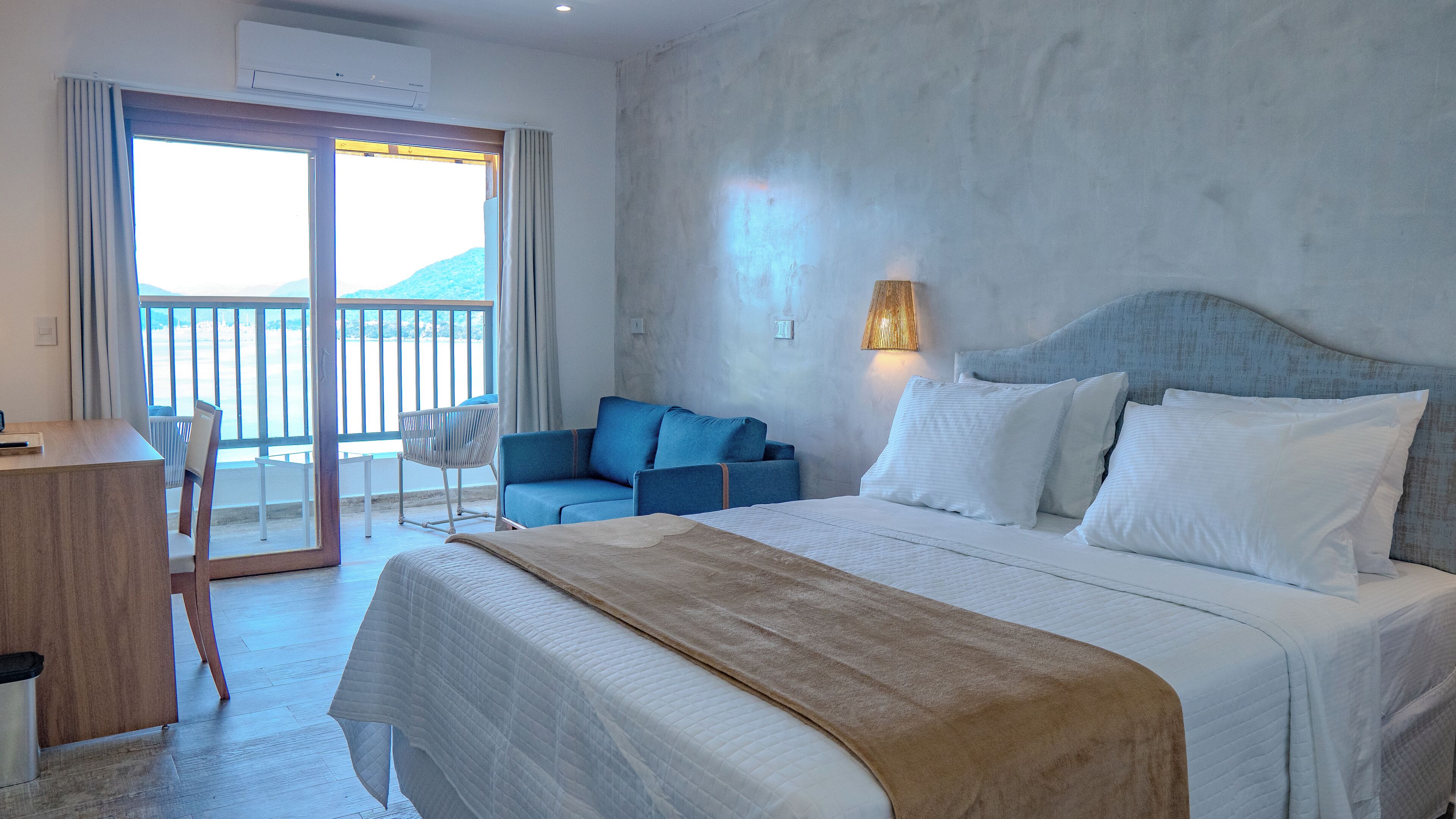 A hotel room with a bed, a desk, a chair, and a balcony with a view of the mountains.