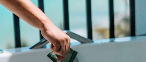 Una mano sostiene una esponja sobre una bañera con vista a un cuerpo de agua a través de una ventana con barras negras.