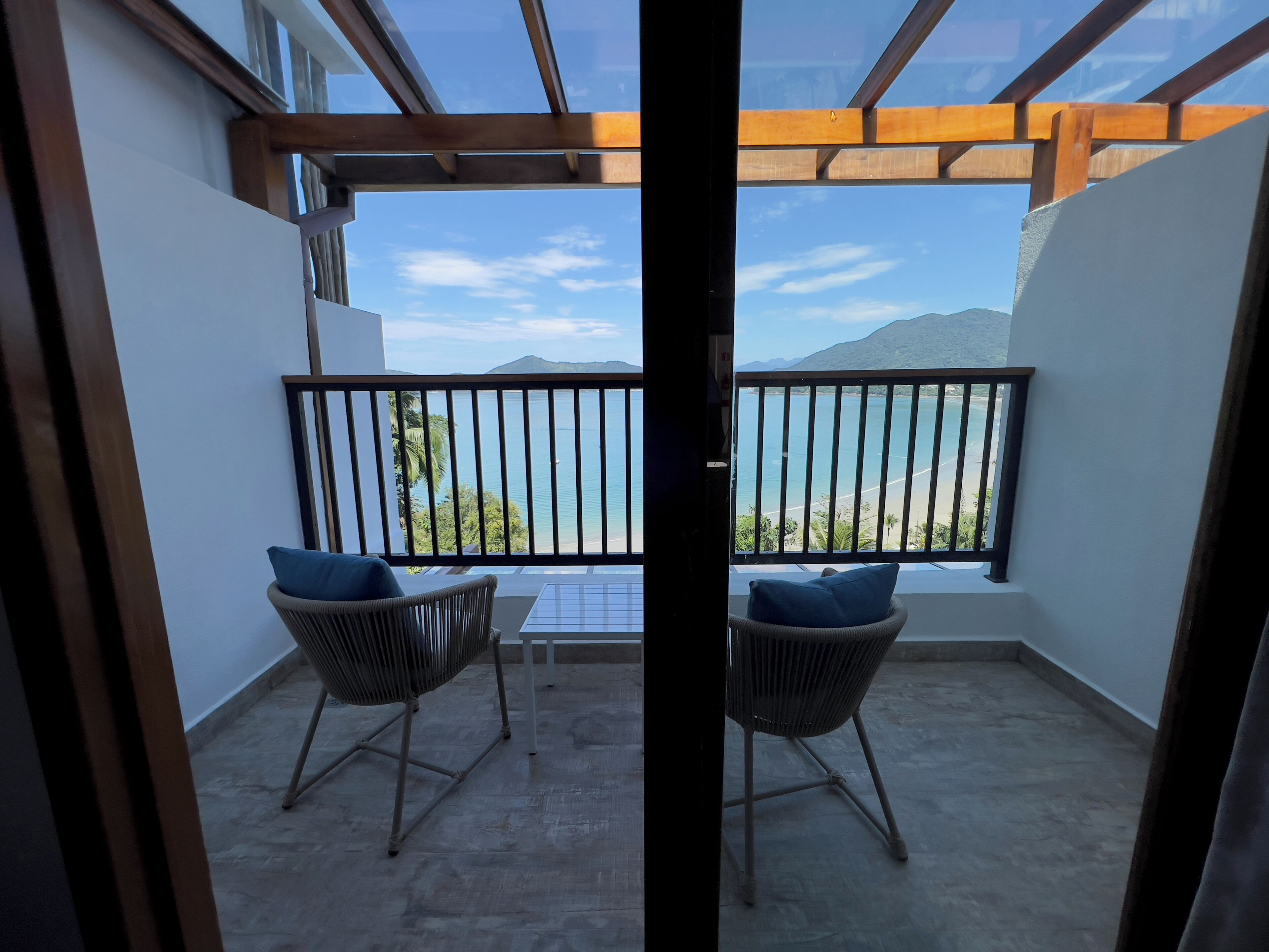 A balcony with two wicker chairs and a table, overlooking a beach and mountains.