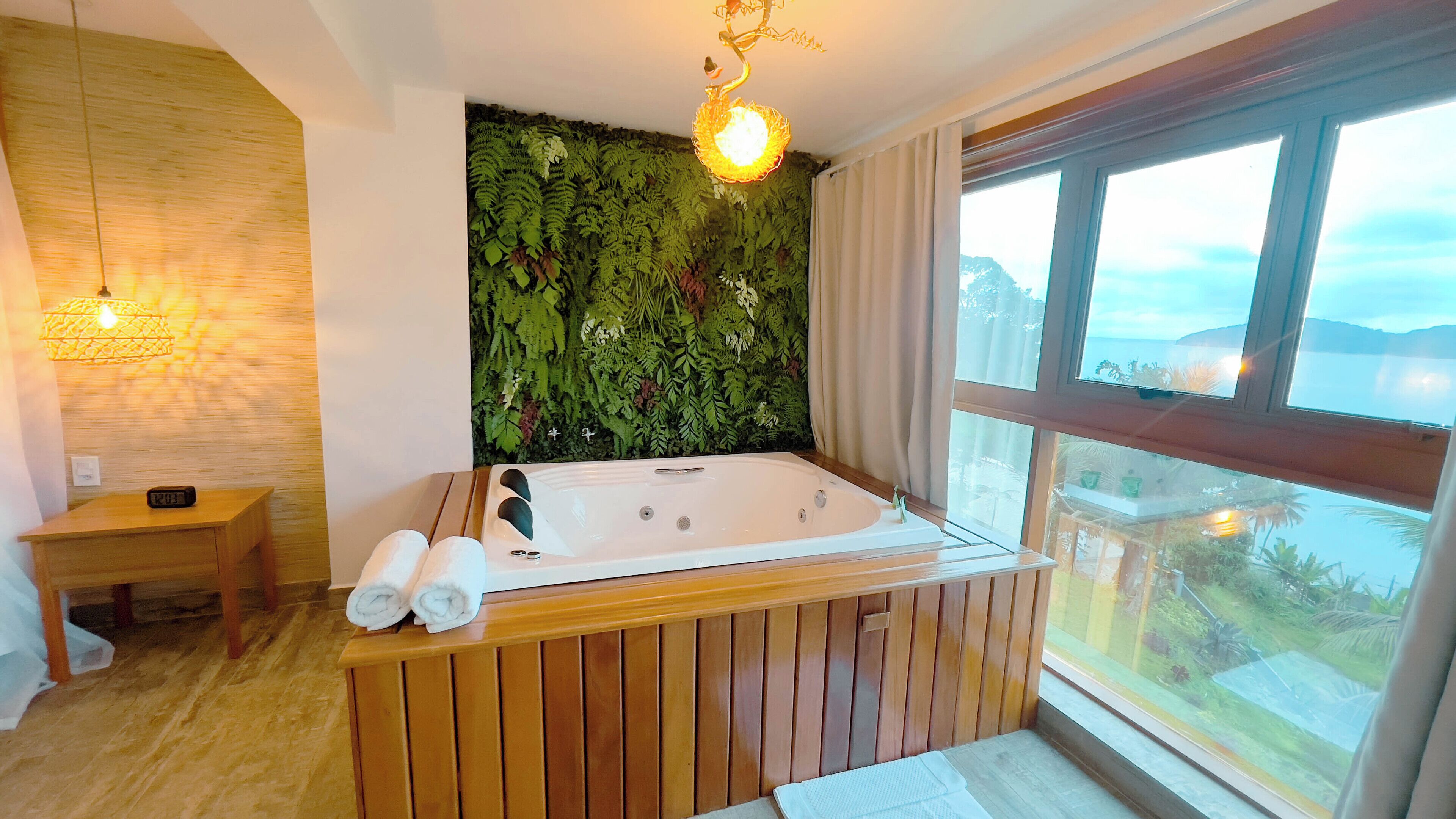 Une salle de bain moderne avec une baignoire en bois, un mur végétal, une table de chevet en bois et une vue sur la mer grâce à de grandes fenêtres.