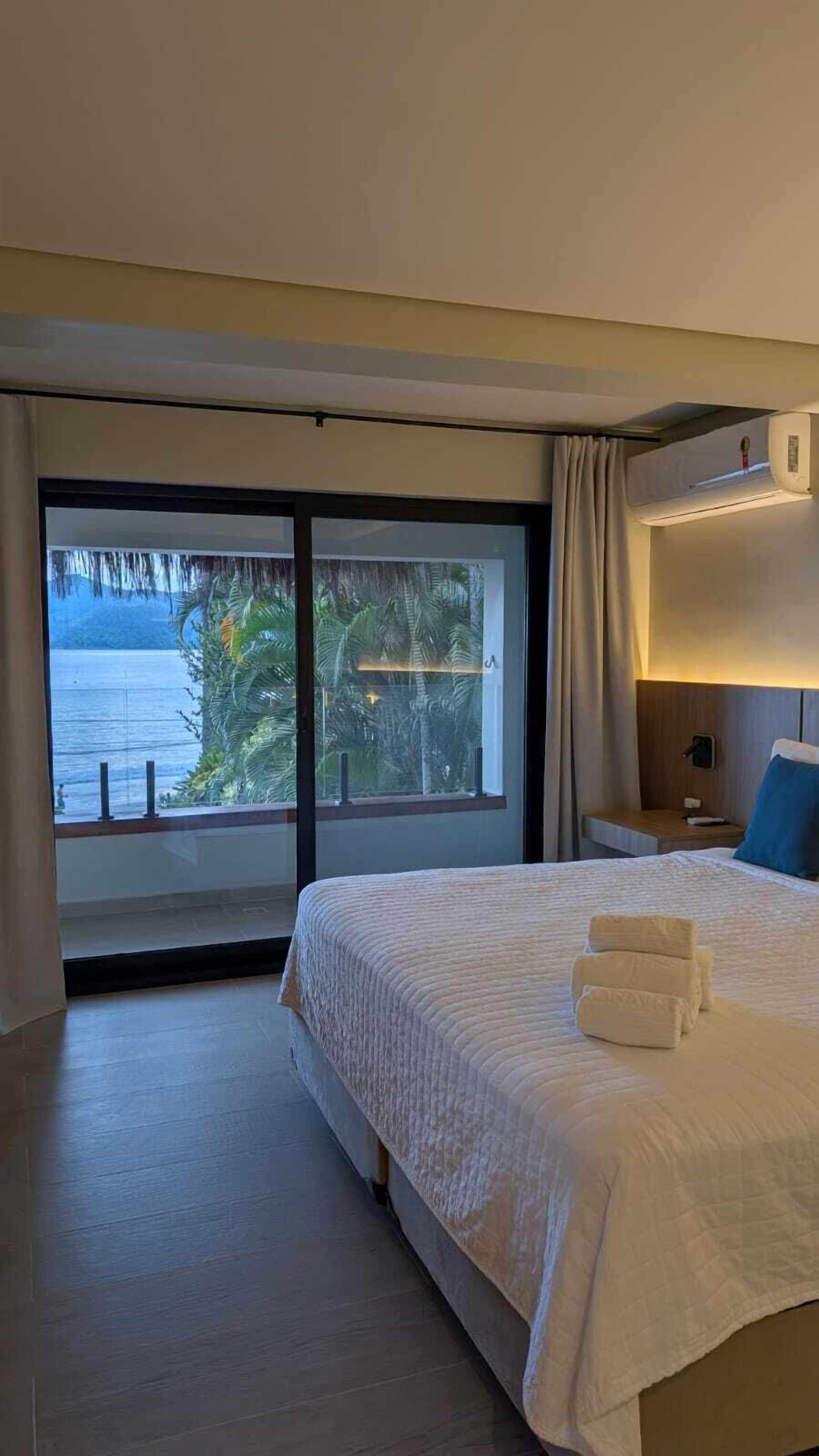 Ein Hotelzimmer mit einem großen Bett, einem Nachttisch, einer Wand-Klimaanlage und Meerblick durch eine Schiebetür.