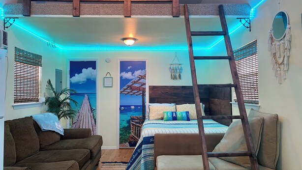 Ein gemütliches Schlafzimmer mit einem Schlafplatz in der Schlaffküche, einer Holzleiter, einer Hängematte und Meerblick durch eine Tür.