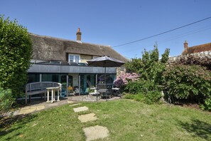 A thatched-roof building with a patio area featuring a bench, table, and umbrella. - Stable Cottage (Bridport)