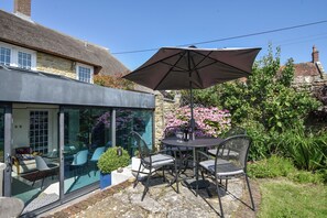 A glass-enclosed patio with a table and chairs, a large umbrella, and a garden with pink flowers. - Stable Cottage (Bridport)