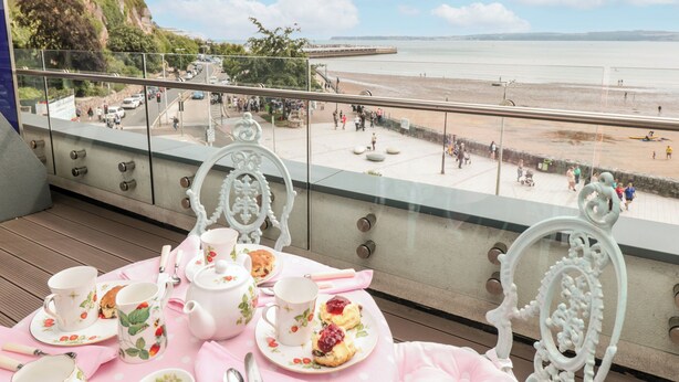 A balcony with a table set for tea, overlooking a beach and promenade.