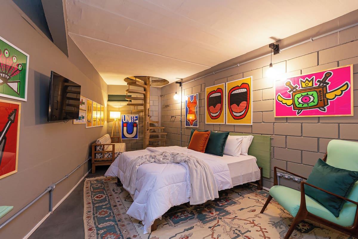 Ein Schlafzimmer mit einem Bett, einem Sessel, einer Wand mit farbenfroher Kunst und einer Wendeltreppe.