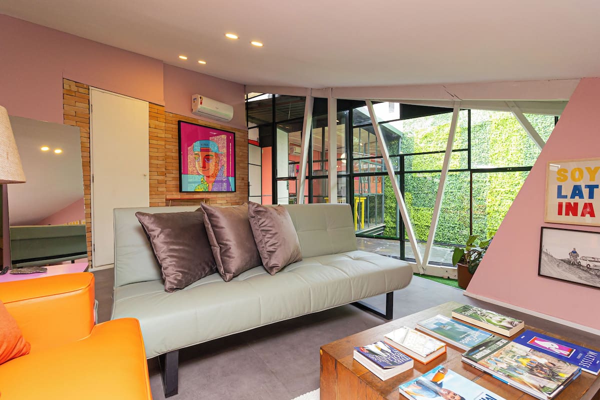 Ein modernes Wohnzimmer mit einem hellgrauen Sofa, einem Couchtisch mit Zeitschriften, einer Lampe und einem großen Fenster mit Blick auf Grünflächen.