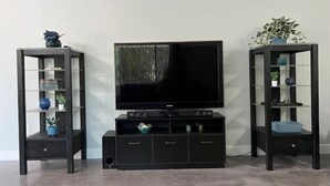 A living room with a flat-screen TV on a black stand, flanked by two black shelving units with glass doors, each containing decorative items and plants. - Modern Universal Orlando (Orlando)