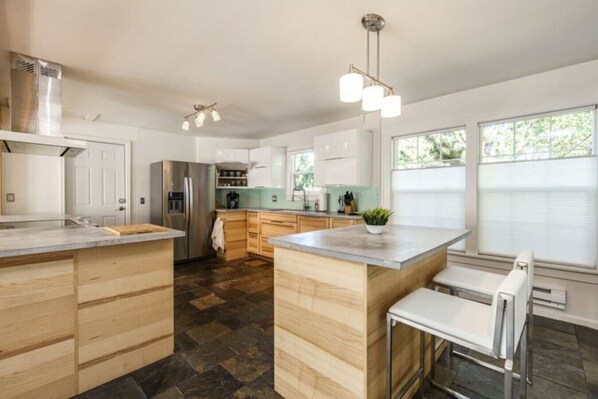 Una cocina moderna con una isla central, muebles de madera, un refrigerador de acero inoxidable y una zona de comedor con una silla blanca.