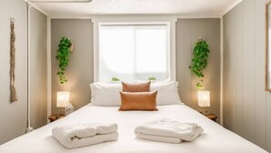 A neatly made bed with white linens, two brown pillows, and two folded towels. There are two bedside tables with lamps, and green plants hanging on the walls.