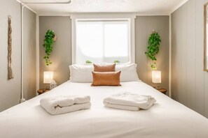 A neatly made bed with white linens, two brown pillows, and two folded towels. There are two bedside tables with lamps, and green plants hanging on the walls.
