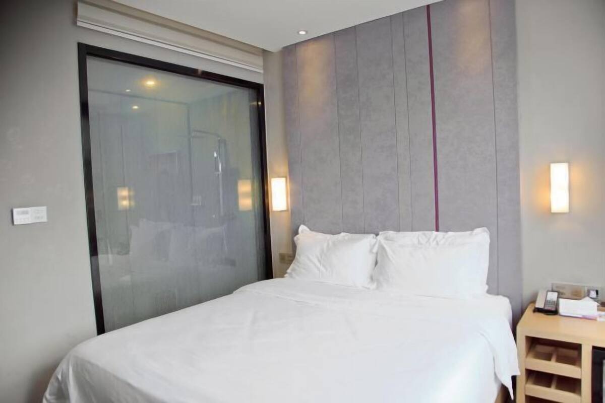 A modern hotel room with a large bed, a glass door leading to a bathroom, and a wooden nightstand with a lamp.