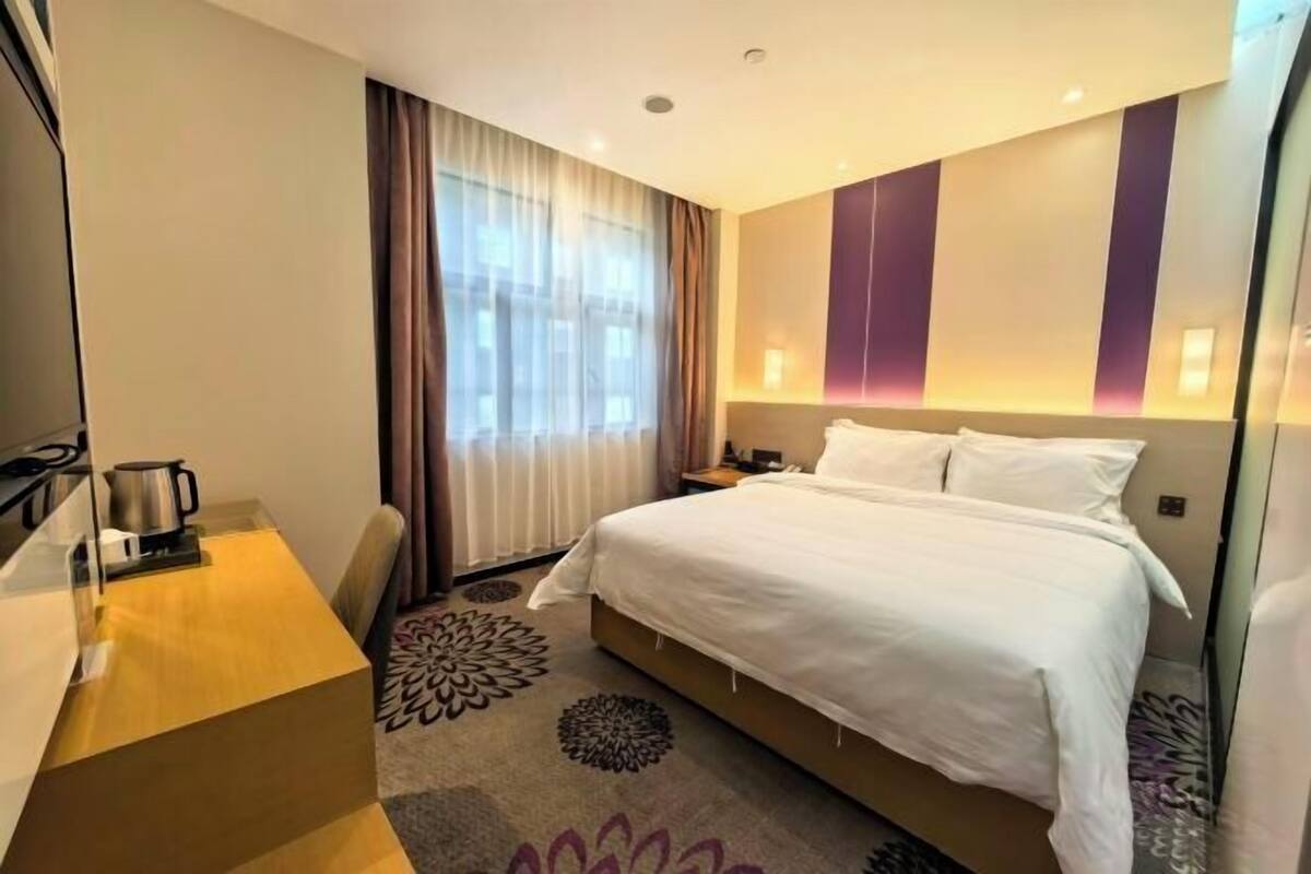 A hotel room with a bed, a desk, a chair, a window with curtains, and a patterned carpet.