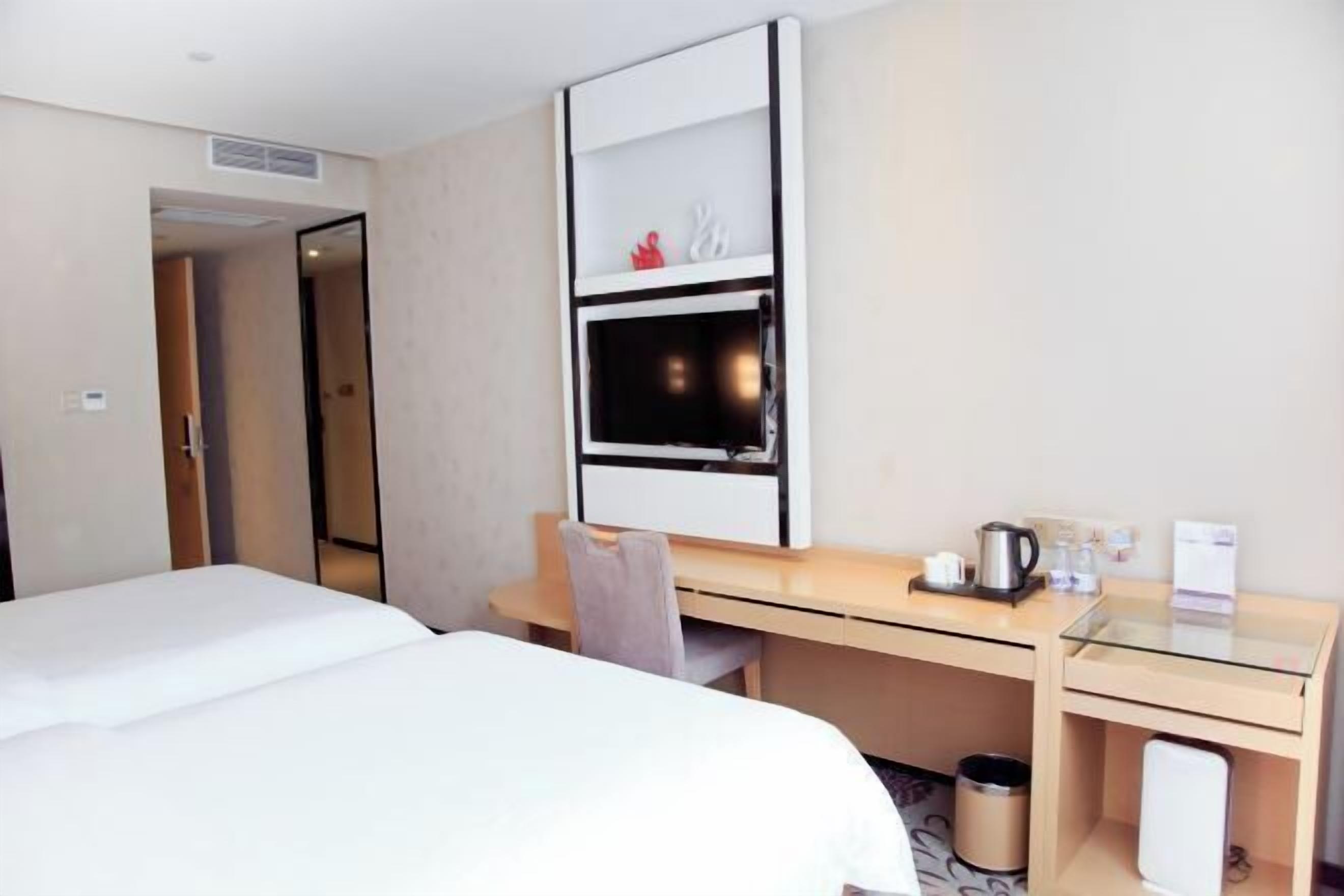 A hotel room with a bed, a desk, a chair, a mirror, a shelf with decorative items, and a trash bin.