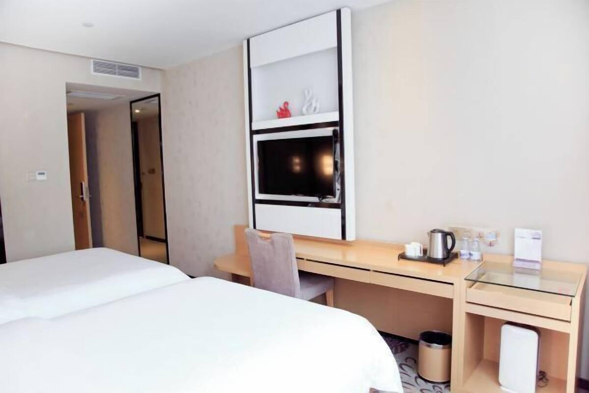 A hotel room with a bed, a desk, a chair, a mirror, a shelf with decorative items, and a trash bin.