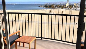 Un balcón con una mesa y silla de madera, con vistas a una playa de arena, palmeras y un cielo azul despejado.