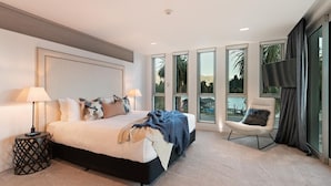 Quarto moderno com cama grande, duas luminárias de cabeceira, uma cadeira e janelas amplas com vista para o exterior.