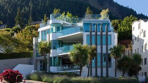 Um edifício moderno de vários andares com varandas, rodeado por vegetação e com montanhas ao fundo.