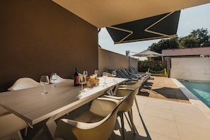Uma área de refeições ao ar livre com piscina, incluindo uma mesa comprida, cadeiras e guarda-sóis.