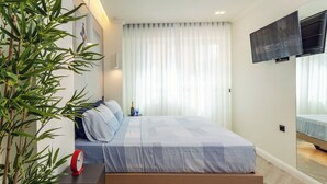 A modern bedroom with a bed, a bedside table with a clock, a TV mounted on the wall, and a large window with sheer curtains.