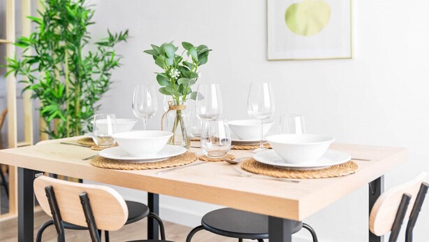 Uma mesa de jantar posta com pratos, tigelas e copos de vinho brancos, acompanhada por uma pequena planta e um quadro na parede.