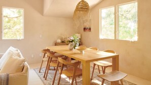 Un área de comedor con una mesa y sillas de madera, una lámpara colgante y ventanas que ofrecen vistas al verdor.