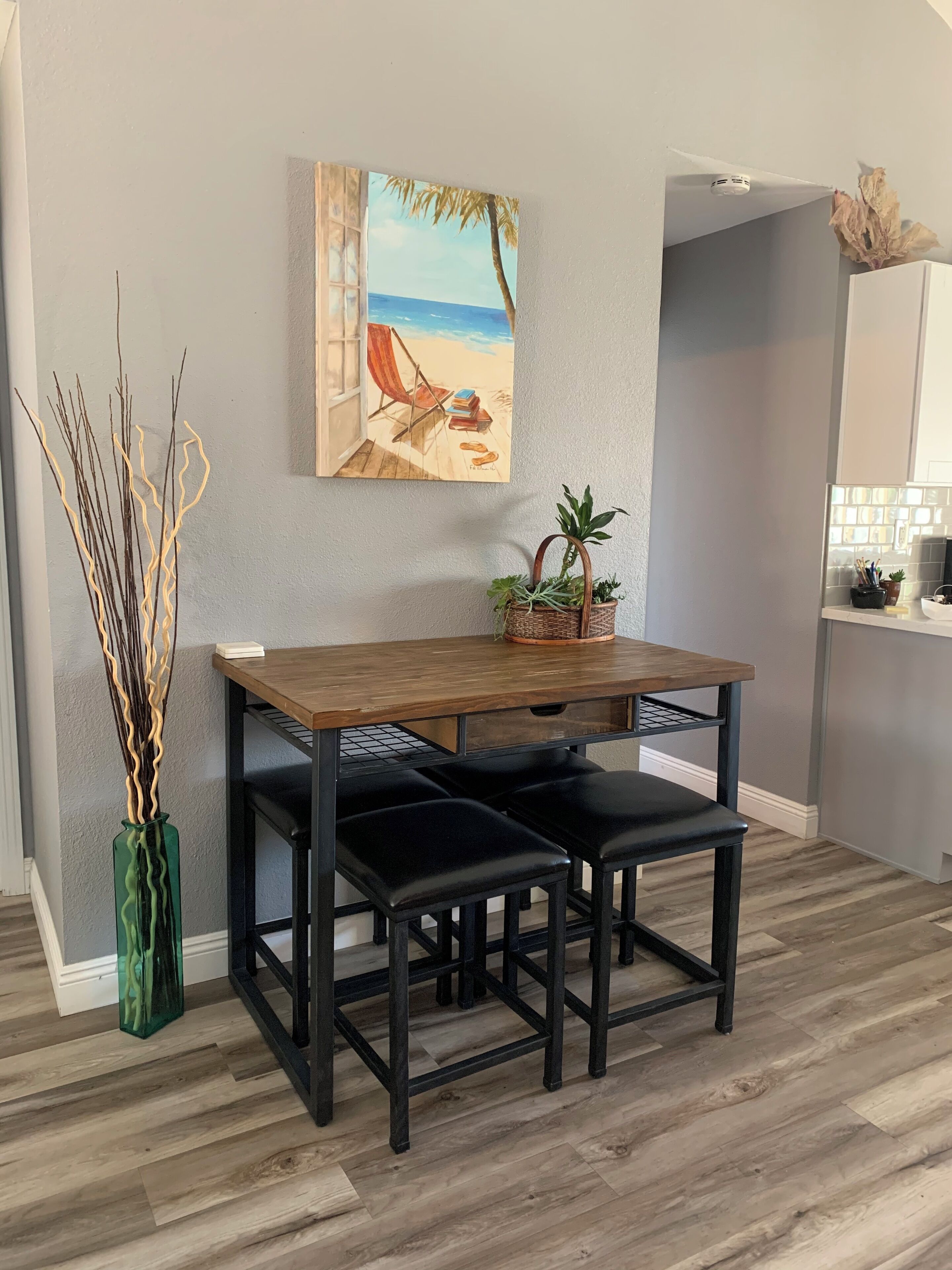 A dining area with a wooden table and black chairs, a painting of a beach scene, a green vase with branches, and a small potted plant on the table. - COZY UCSD/La Jolla Condo 2bd, 10-15 min from just about anywhere in the city (San Diego)